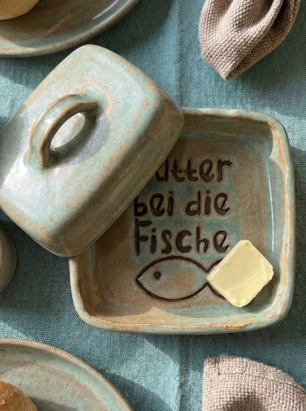 Handgemachte Keramik-Butterdose in Blaugrün und Hellbraun glasiert. Im Boden ist der Spruch 'Butter bei die Fische' mit einem Fisch tief eingestempelt. Eine kreative Töpfer-Idee.