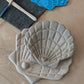 Zwei übereinandergelegte Muschelschalen aus Ton mit detailreicher Relief-Struktur, im Hintergrund die blaue Prägeplatte und Werkzeug.