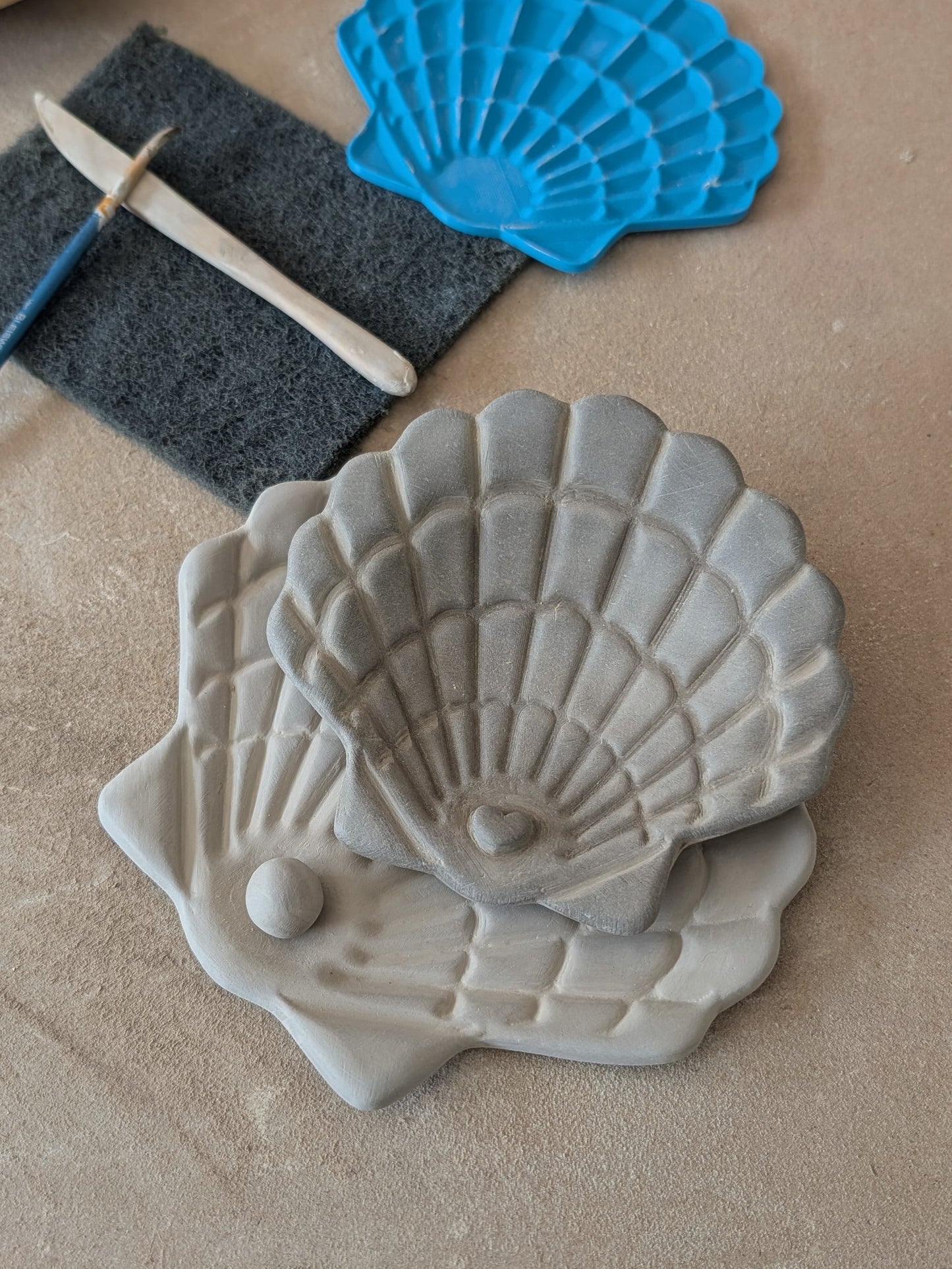 Zwei übereinandergelegte Muschelschalen aus Ton mit detailreicher Relief-Struktur, im Hintergrund die blaue Prägeplatte und Werkzeug.