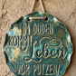 Ein fertig glasiertes, gewelltes Keramikschild mit dem eingestempelten Spruch 'Im Duden kommt Leben vor Putzen!'. Die glänzende blaugrüne Glasur betont das tiefe Relief der Schrift auf hellem Ton. Das Schild hängt an einer rustikalen Kordel an einem Nagel vor einer Lehmwand – ein ideales Anwendungsbeispiel für MinseArt Keramikstempel.