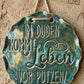 Ein fertig glasiertes, gewelltes Keramikschild mit dem eingestempelten Spruch 'Im Duden kommt Leben vor Putzen!'. Die glänzende blaugrüne Glasur betont das tiefe Relief der Schrift auf hellem Ton. Das Schild hängt an einer rustikalen Kordel an einem Nagel vor einer Lehmwand – ein ideales Anwendungsbeispiel für MinseArt Keramikstempel.