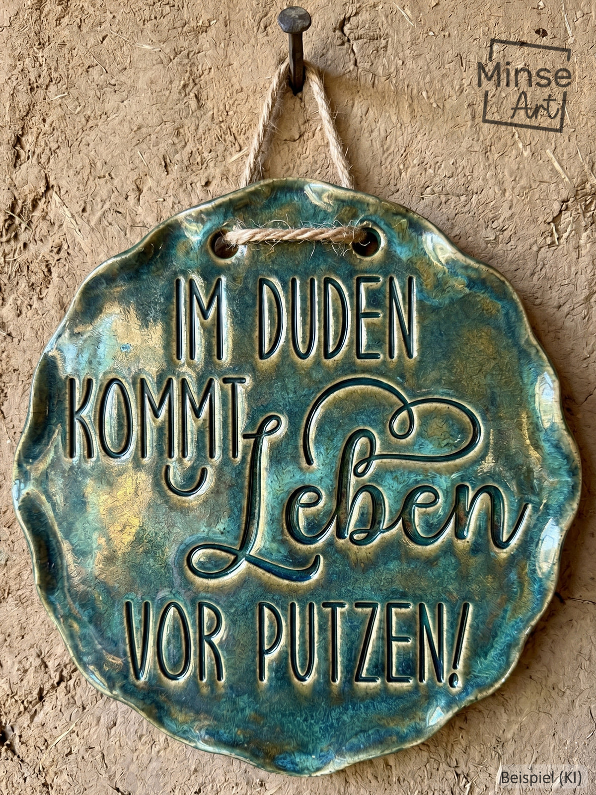 Ein fertig glasiertes, gewelltes Keramikschild mit dem eingestempelten Spruch 'Im Duden kommt Leben vor Putzen!'. Die glänzende blaugrüne Glasur betont das tiefe Relief der Schrift auf hellem Ton. Das Schild hängt an einer rustikalen Kordel an einem Nagel vor einer Lehmwand – ein ideales Anwendungsbeispiel für MinseArt Keramikstempel.