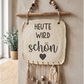 Getöpfertes Keramik-Wandschild im Boho-Stil mit eingestempeltem Spruch Heute wird schön, Herz-Detail, Holzaufhängung und Makramee-Verzierungen.