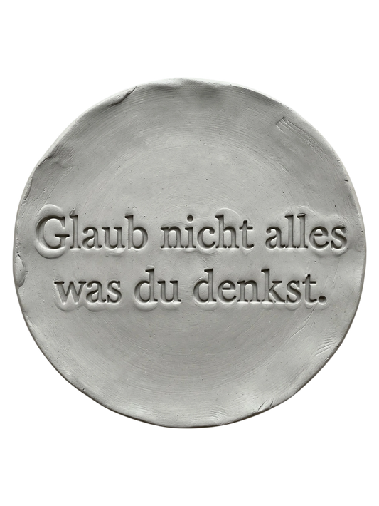 Präziser Stempelabdruck 'Glaub nicht alles was du denkst' in hellem Ton. Sauberes, tiefes Relief auf einer runden Tonplatte, erstellt mit einem MinseArt Textstempel.