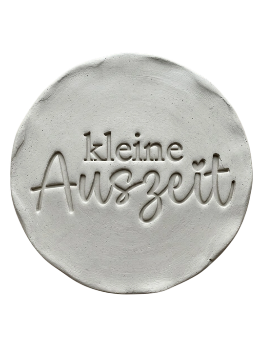 Präziser Stempelabdruck 'kleine Auszeit' in hellem Ton. Das saubere Relief kombiniert eine moderne Serifenschrift mit eleganter Schreibschrift auf einer runden Tonplatte.