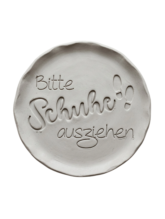 Präziser Stempelabdruck 'Bitte Schuhe ausziehen' in hellem Ton mit kleinen Fußabdruck-Details. Sauberes Relief für die Gestaltung von Flur-Deko.