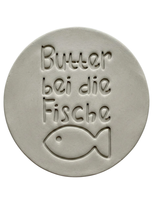 Detailreicher Stempelabdruck des Spruchs 'Butter bei die Fische' mit einem Fisch-Motiv in hellem Ton. Präzise Konturen für handgemachte Töpferprojekte.