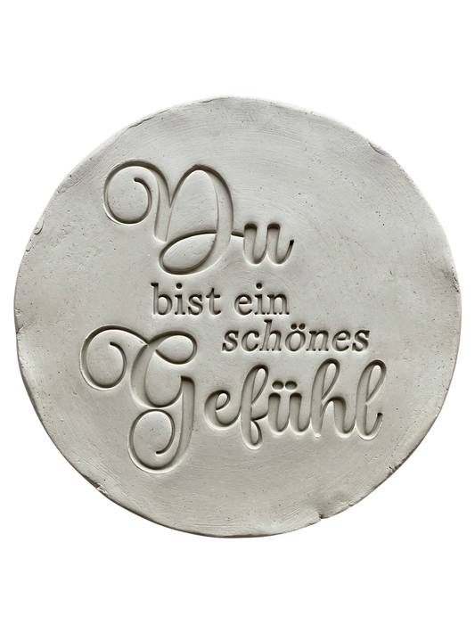 Präziser Stempelabdruck des Spruchs 'Du bist ein schönes Gefühl' in hellem Ton. Die elegante Kombination aus schwungvoller Schreibschrift und klassischer Serifenschrift erzeugt ein tiefes Relief auf der runden Tonplatte.