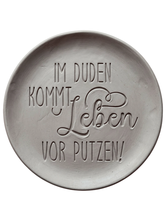Witziger Stempelabdruck 'Im Duden kommt Leben vor Putzen' in hellem Ton. Sauberes Relief mit einer Kombination aus moderner Druckschrift und schwungvoller Schreibschrift.