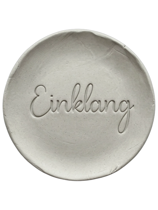 Präziser Stempelabdruck des Wortes 'Einklang' in eleganter Schreibschrift auf einer runden Tonplatte. Filigranes Relief für spirituelle Keramik, gestaltet mit einem MinseArt Keramikstempel.