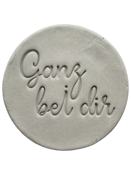 Filigraner Stempelabdruck in eleganter Handschrift mit dem Spruch 'Ganz bei dir' auf einer runden Tonplatte. Sauberes Relief-Ergebnis für meditative Keramik, gestaltet mit einem MinseArt Keramikstempel.
