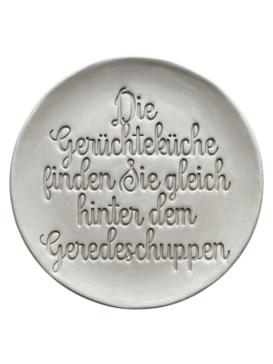 Präziser Stempelabdruck des Spruchs 'Die Gerüchteküche finden Sie gleich hinter dem Geredeschuppen' in hellem Ton. Sauberes Relief in eleganter Schreibschrift, erstellt mit einem MinseArt Keramikstempel.