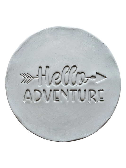 Präziser Abdruck im hellen Ton mit dem Spruch 'Hello Adventure'. Eine kreative Töpfer-Idee für Reisetagebücher aus Ton, Wandteller oder Geschenke zum Abenteuer-Start.