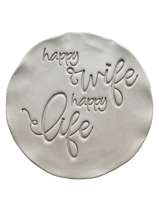 Präziser Stempelabdruck 'happy wife happy life' in hellem Ton. Die schwungvolle, moderne Schreibschrift erzeugt ein sauberes, tiefes Relief auf der runden Tonplatte.
