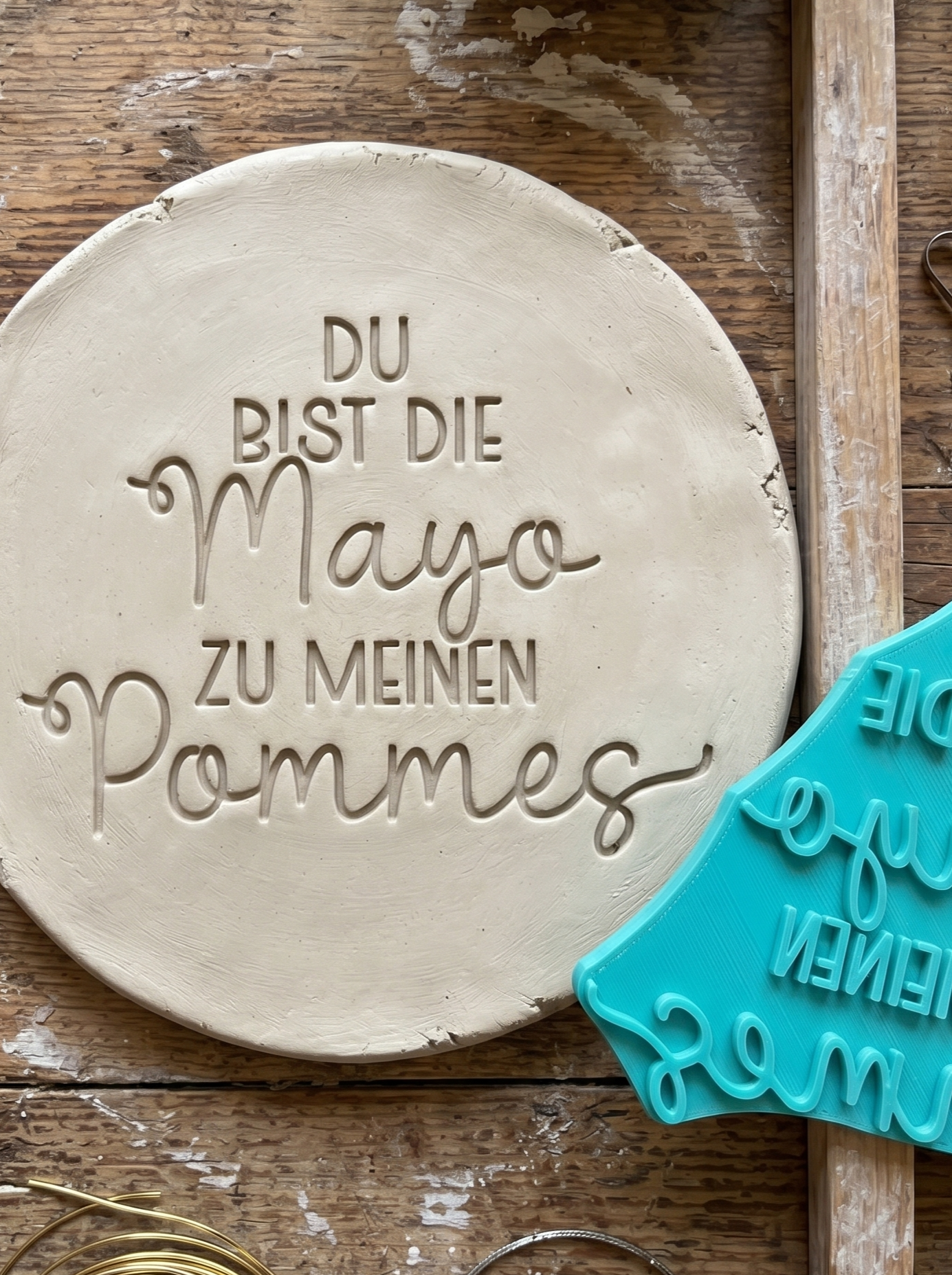 Ein sauberer Stempelabdruck des Spruchs 'Du bist die Mayo zu meinen Pommes' in einer runden Tonplatte. Daneben liegt der türkisfarbene MinseArt Keramikstempel auf einem rustikalen Holztisch zusammen mit Töpferwerkzeug und Abstandshaltern für die perfekte Plattendicke.