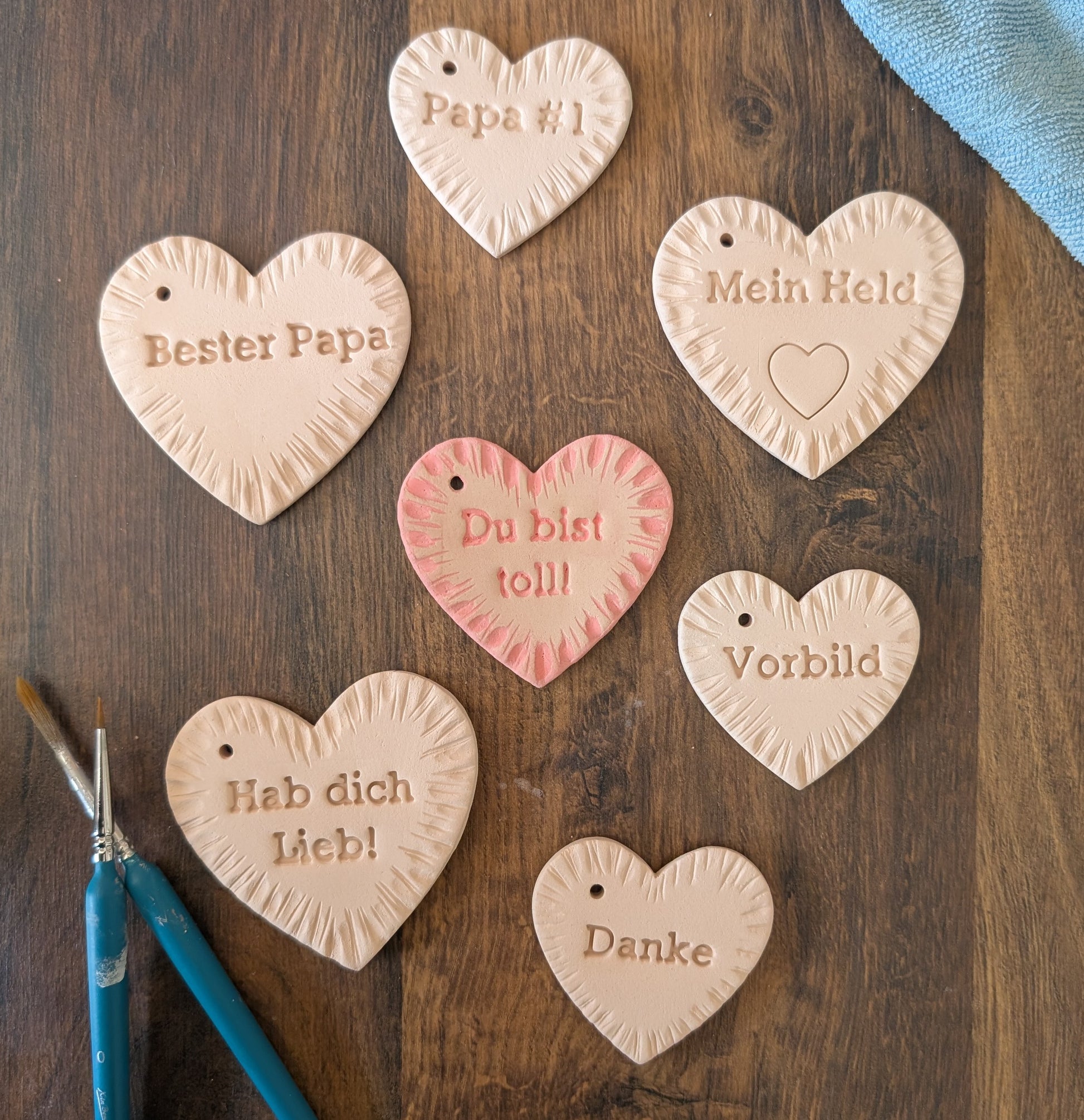 Sieben handgetöpferte Herzanhänger aus Terracotta-Ton mit verschiedenen Papa-Botschaften wie 'Bester Papa', 'Mein Held' und 'Danke'. Kreative DIY-Geschenkidee zum Vatertag.