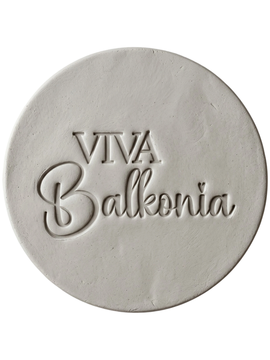 Präziser Stempelabdruck des Schriftzugs 'VIVA Balkonia' in hellem Ton. Das saubere, tiefe Relief auf der runden Tonplatte zeigt die harmonische Kombination aus klarer Druckschrift und verspielter Schreibschrift mit einem kleinen Herz-Detail über dem i. Ein perfektes Töpfer-Ergebnis mit einem MinseArt Keramikstempel.