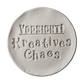 Sauberer Stempelabdruck 'Vorsicht! Kreatives Chaos' in hellem Ton. Die verspielte Typografie mit dekorativen Serifen erzeugt ein präzises Relief auf der runden Tonplatte.