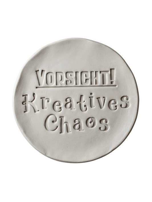 Sauberer Stempelabdruck 'Vorsicht! Kreatives Chaos' in hellem Ton. Die verspielte Typografie mit dekorativen Serifen erzeugt ein präzises Relief auf der runden Tonplatte.