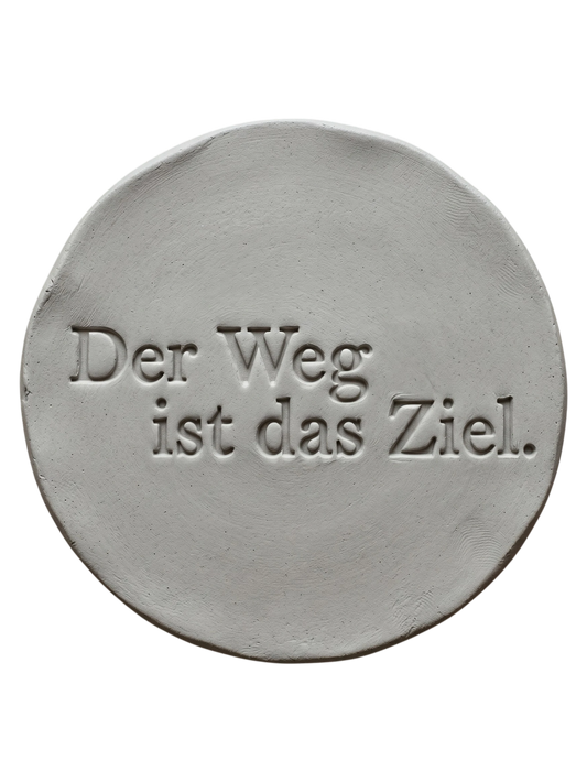 Präziser Stempelabdruck 'Der Weg ist das Ziel' in hellem Ton. Sauberes, tiefes Relief auf einer runden Tonplatte, erstellt mit einem MinseArt Textstempel