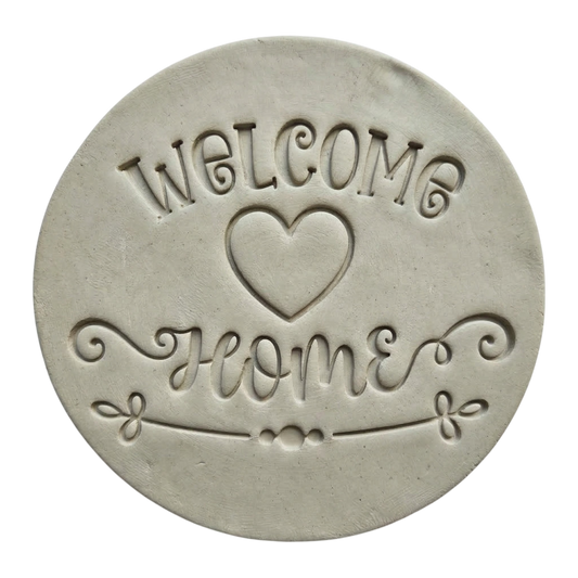 Präziser Stempelabdruck 'Welcome Home' mit Herz-Motiv in hellem Ton. Das saubere Relief kombiniert verspielte Druckschrift mit eleganter Schreibschrift auf einer runden Tonplatte.