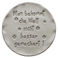 Präziser Stempelabdruck des Spruchs 'Man bekommt die Welt nicht besser gemeckert!' in hellem Ton mit kleinen Blumen-Details. Ein sauberes Relief auf einer runden Tonplatte, erstellt mit einem MinseArt Keramikstempel.