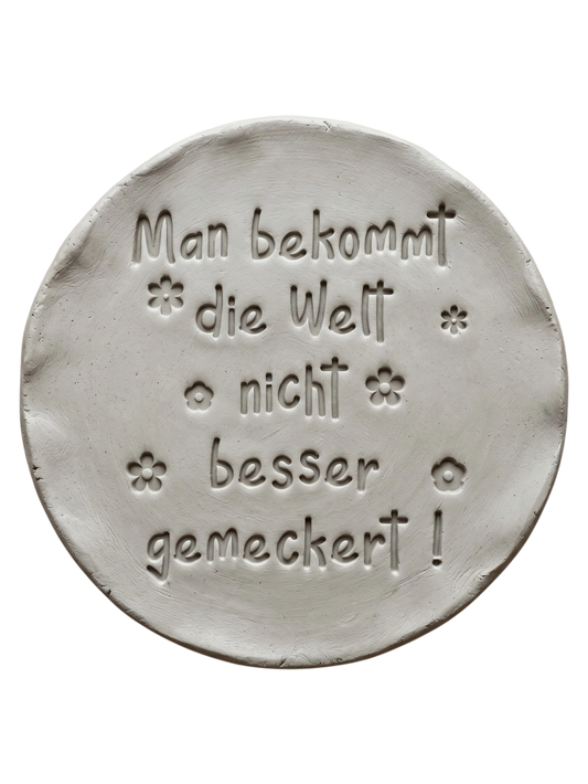 Präziser Stempelabdruck des Spruchs 'Man bekommt die Welt nicht besser gemeckert!' in hellem Ton mit kleinen Blumen-Details. Ein sauberes Relief auf einer runden Tonplatte, erstellt mit einem MinseArt Keramikstempel.