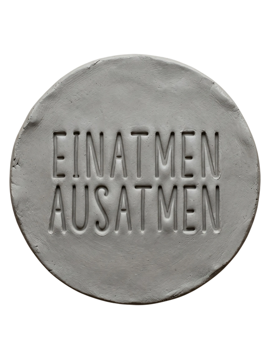 Sauberer Stempelabdruck 'Einatmen Ausatmen' in hellem Ton. Das tiefe Relief der klaren Typografie sorgt für ein minimalistisches Design auf der runden Tonplatte.