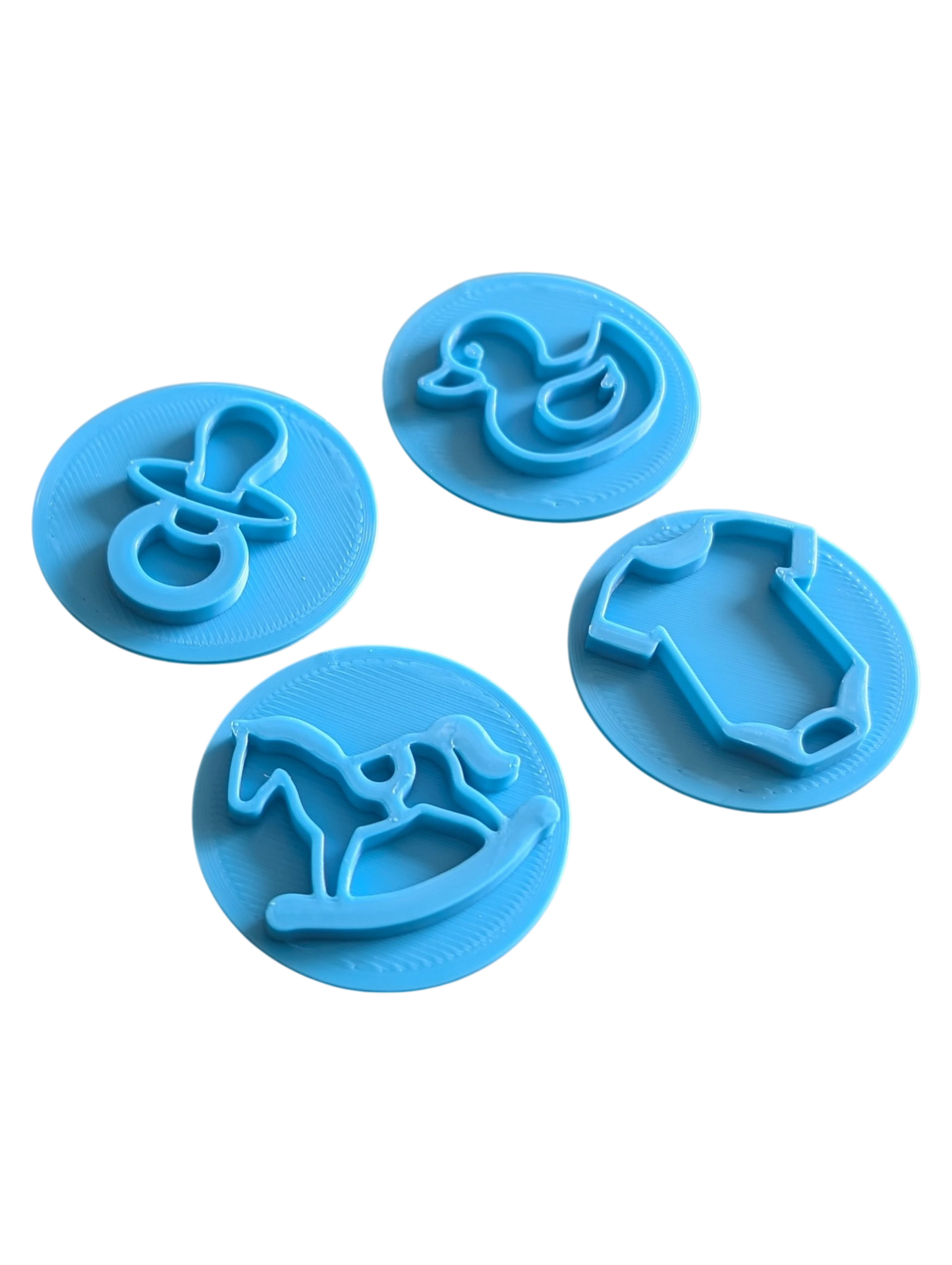 Draufsicht auf das vierteilige Keramikstempel-Set für Ton mit süßen Baby-Motiven. Die runden, gedämpft türkisfarbenen Prägestempel zeigen detailreich einen Schnuller, eine Ente, ein Schaukelpferd und einen Body.