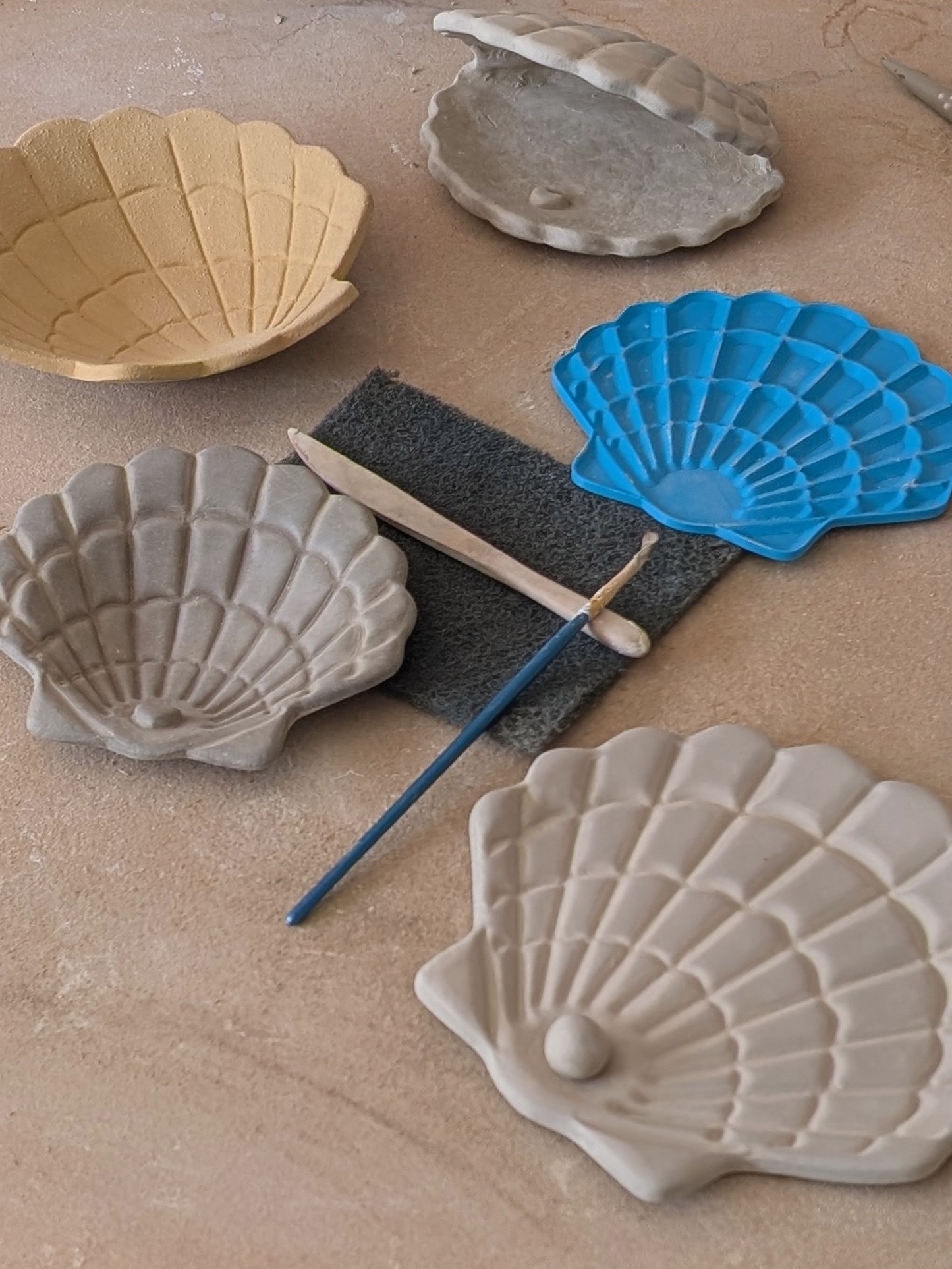 Muschel-Prägeplatte aus blauem Material drapiert neben fertig geformten Muschelschalen und Töpferzubehör.