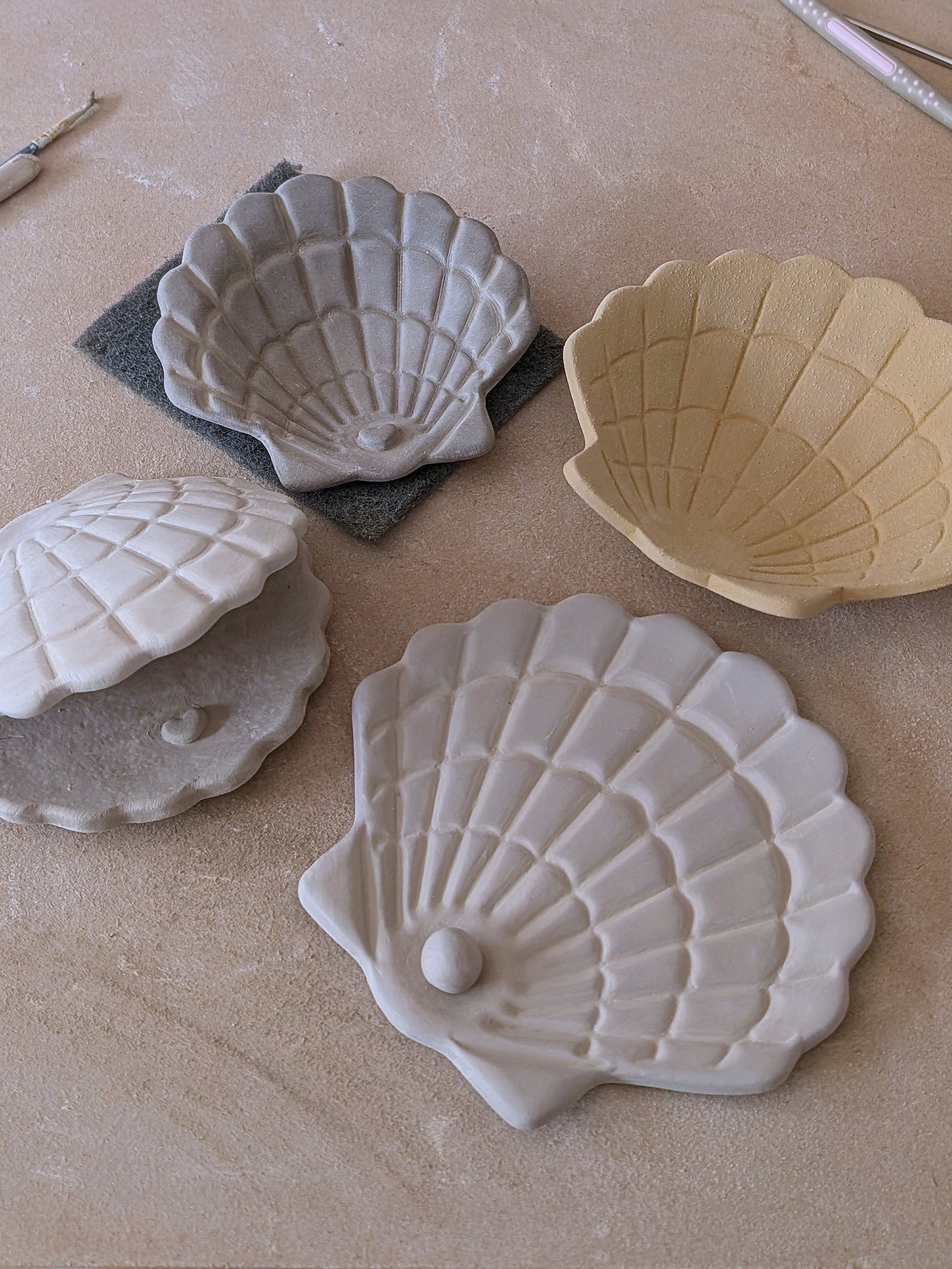 Detailaufnahme handgemachter Muschelschalen aus Ton in verschiedenen Farben mit feiner Reliefstruktur und eingearbeiteter Perle.