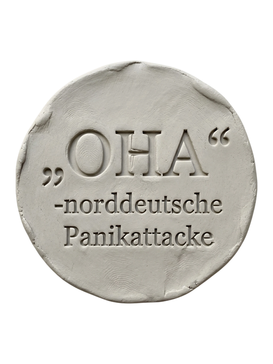 Präziser Stempelabdruck des witzigen Spruchs 'OHA - norddeutsche Panikattacke' in hellem Ton. Sauberes, tiefes Relief mit klarer Typografie auf einer runden Tonplatte.