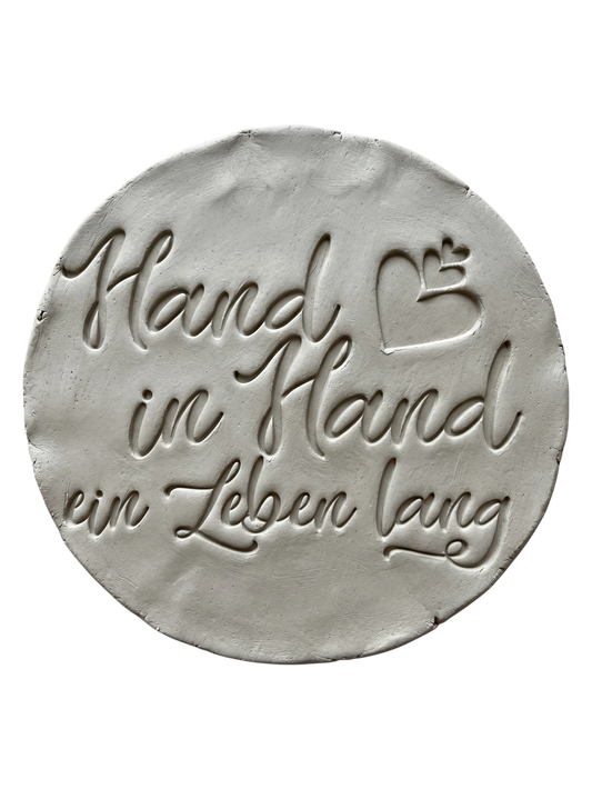Präziser Stempelabdruck des Spruchs 'Hand in Hand ein Leben lang' in hellem Ton. Detailreiches, sauberes Relief in eleganter Schreibschrift mit Herzen auf einer runden Tonplatte. Handgefertigt mit einem MinseArt Textstempel.