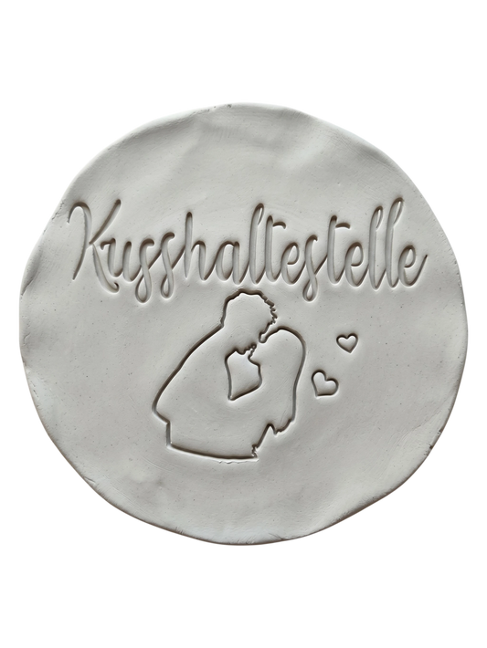 Sauberer und präziser Stempelabdruck des Motivs Kusshaltestelle in hellem Ton. Das detailreiche Relief zeigt ein küssendes Paar und kleine Herzen auf einer runden Tonplatte.