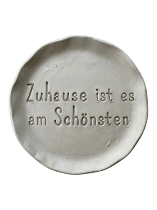 Detailreicher Stempelabdruck des Spruchs Zuhause ist es am Schönsten in hellem Ton. Das saubere Relief auf der runden Tonplatte zeigt präzise Konturen für dekorative Keramikprojekte.