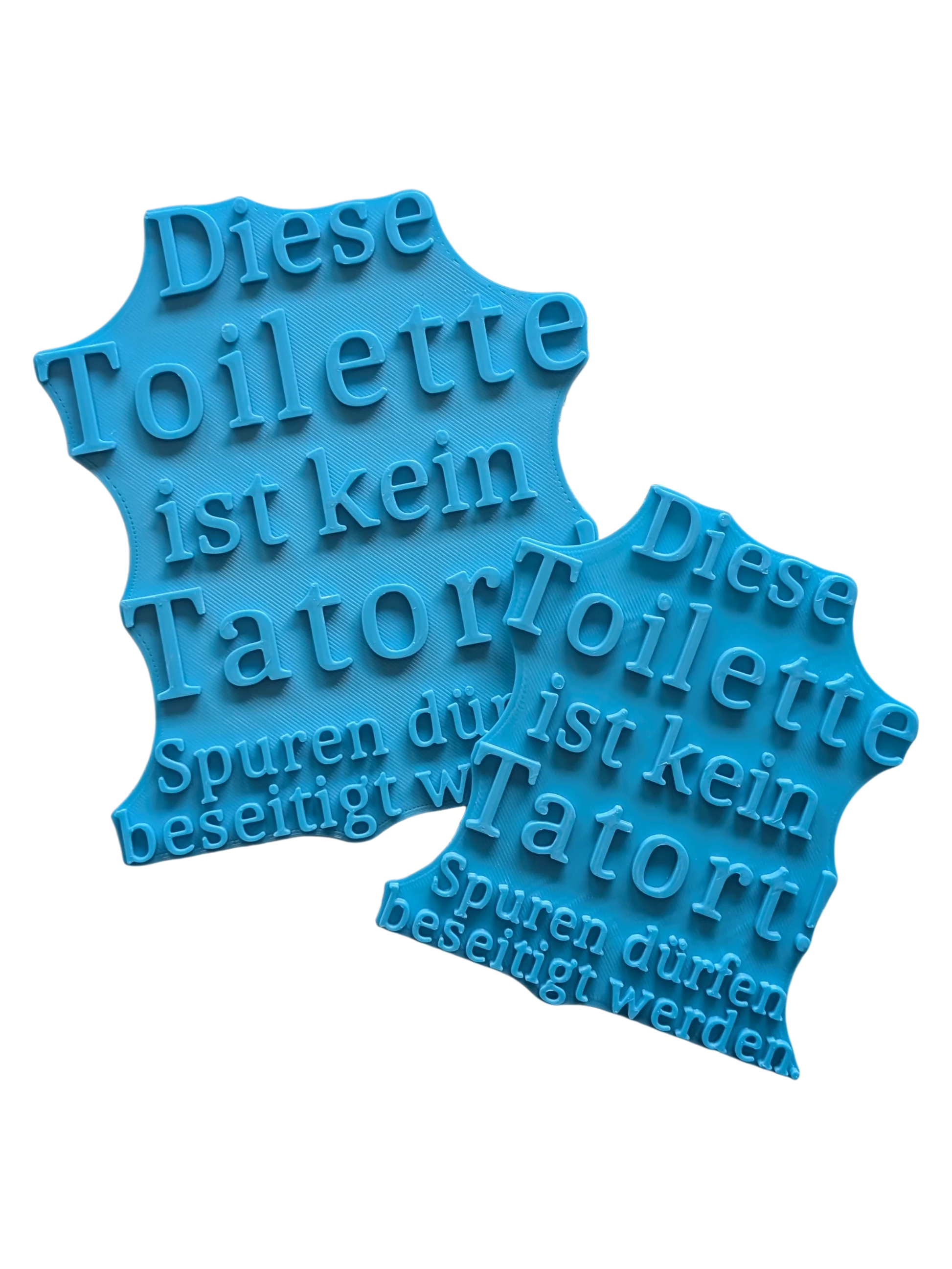 Zwei türkisfarbene Keramikstempel in verschiedenen Größen mit dem humorvollen Spruch 'Diese Toilette ist kein Tatort!'. Ein detailreicher Tonstempel für lustige Badezimmer-Deko und Töpfer-Projekte.