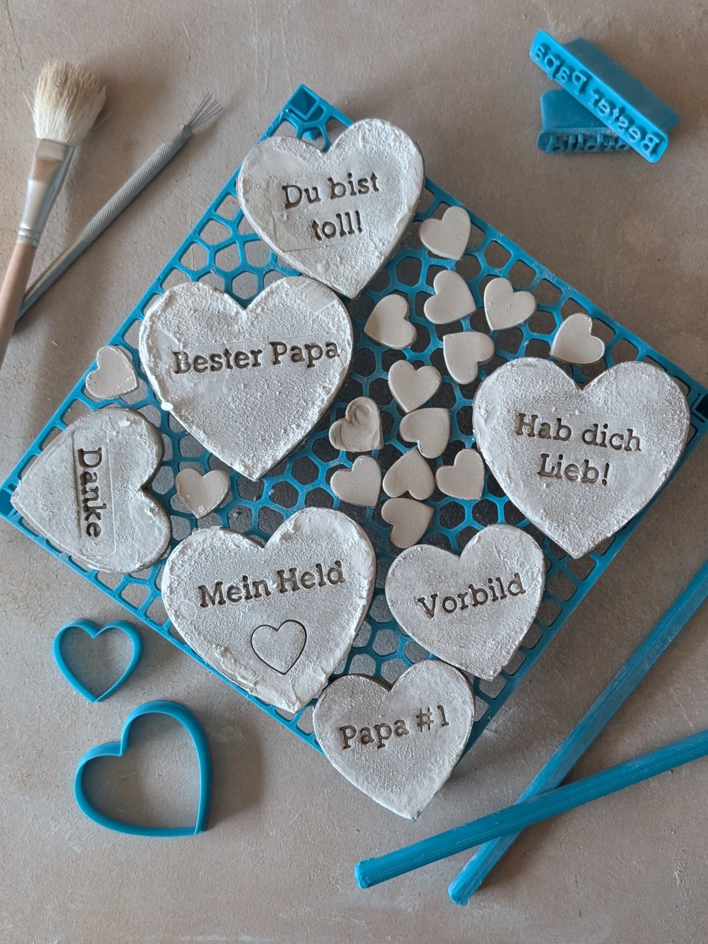 Töpfer-Set in Arbeit: Frische Ton-Herzen mit Stempelprägung auf einer Trocknungsunterlage. Zu sehen sind blaugrüne Keramikstempel und Herz-Ausstecher für die eigene Keramik-Werkstatt.