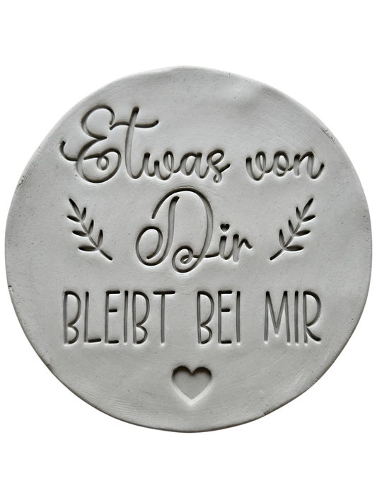 Präziser Stempelabdruck des Textstempels 'Etwas von dir bleibt bei mir' in hellem Ton. Das detailreiche Relief zeigt klare Schrift und florale Elemente perfekt auf einer unglasierten, runden Tonplatte.