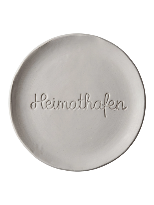 Sauberer Stempelabdruck des Textstempels 'Heimathafen' in hellem, unglasiertem Ton. Die fließende, zusammenhängende Schreibschrift kommt als tiefes Relief auf der runden Tonplatte perfekt zur Geltung.