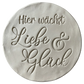 Sauberer Stempelabdruck des Textstempels 'Hier wächst Liebe & Glück' in hellem, unglasiertem Ton. Die elegante Schrift kommt auf der runden Tonplatte wunderschön zur Geltung.
