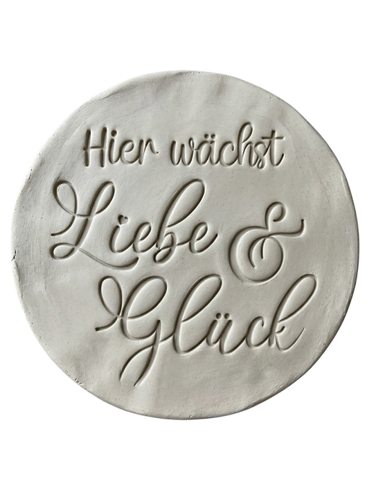 Sauberer Stempelabdruck des Textstempels 'Hier wächst Liebe & Glück' in hellem, unglasiertem Ton. Die elegante Schrift kommt auf der runden Tonplatte wunderschön zur Geltung.