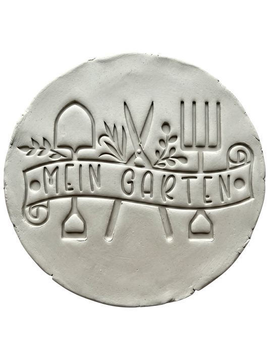 Präziser Stempelabdruck in hellem Ton mit dem Textstempel 'Mein Garten'. Das detailreiche Relief zeigt Gartengeräte und Pflanzenmotive auf einer handgemachten, runden Keramikplatte.