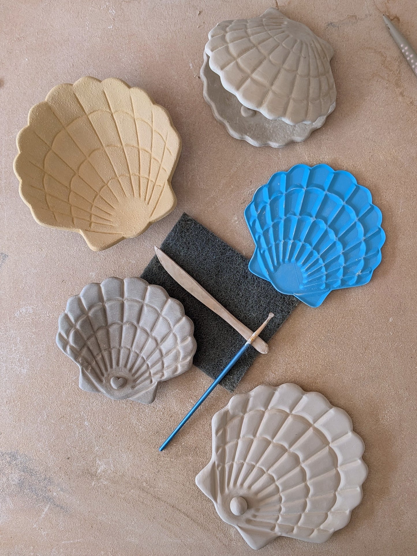 Verschiedene handgeformte Muschelschalen aus Ton neben der blauen Muschel-Prägeplatte von MinseArt und Töpferwerkzeug auf einer Werkbank.